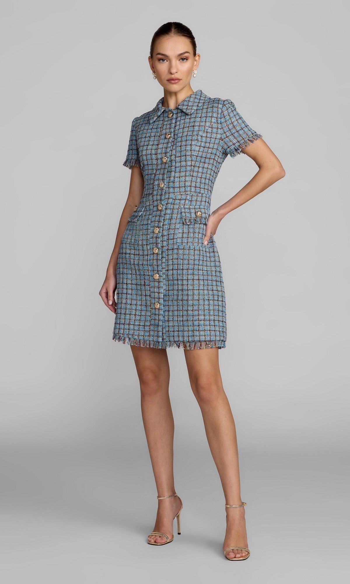 A woman stands with one hand on her hip, modeling the Mac Duggal 21221 Short Boucle Day Dress in Blue Multi. The mini dress features short sleeves, a collared neckline, frayed edges, and gold button detailing. She pairs the look with nude heels against a plain gray background.